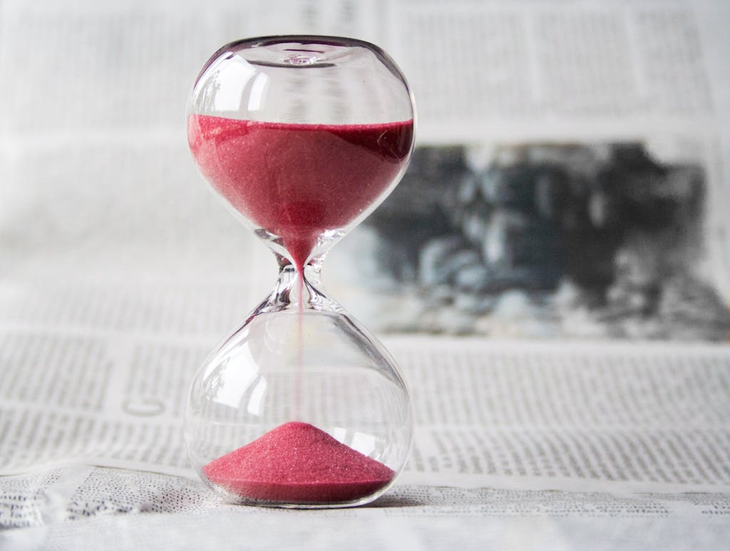 Se Lancer Jeune dans ses Projets : 7 Raisons de ne plus Attendre Close-up of a transparent hourglass with pink sand flowing, placed on a newspaper background.